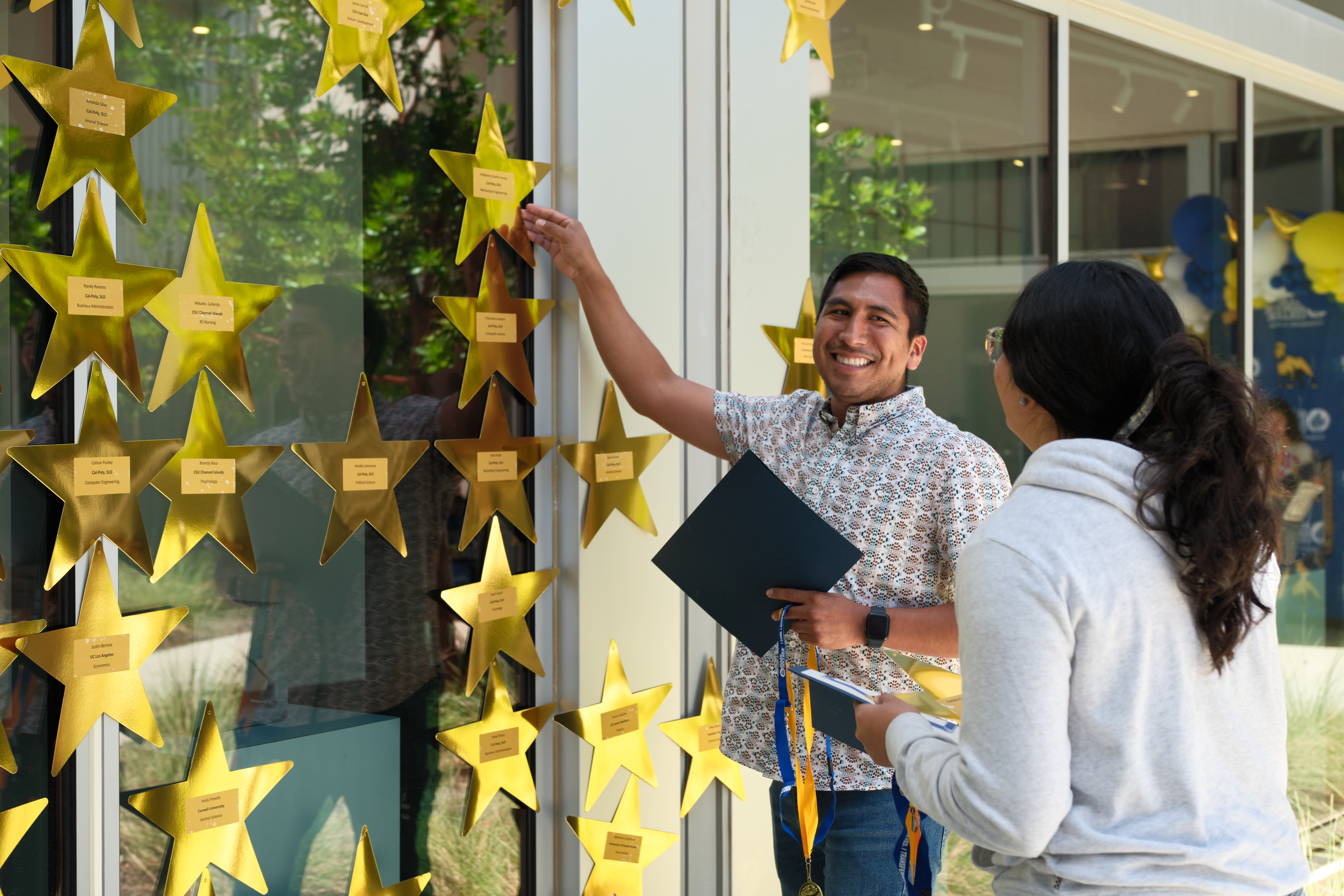 A student smiles while placing a gold star on a window display during Allan Hancock College’s 2026 Transfer Stars Celebration. Another student stands nearby holding papers and a medal ribbon. The display features numerous gold stars recognizing students transferring to four-year colleges and universities.
